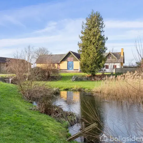 En gul bygning med blå dør ved siden af et stort træ afspejler sig i en dam omgivet af græs og buskads. En lade står til venstre.