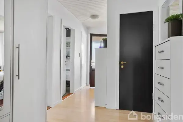 En lang gang har lyse trægulve og hvide vægge. Sort dør med gyldent håndtag til højre, hvide skabe og grøn plante ovenpå.