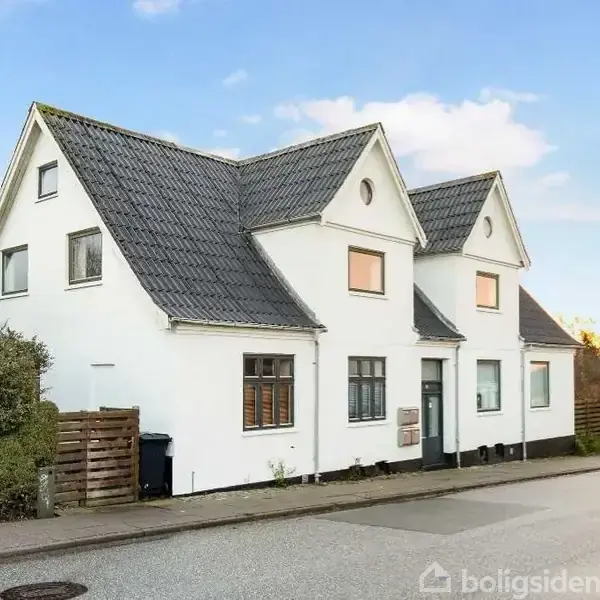 Hvidt hus med to skråtage, står stille langs en villavej. Omkring er der grønne buske og andre huse i baggrunden, under en blå himmel med få skyer.
