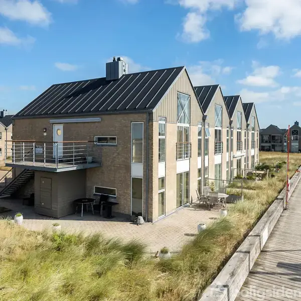 Lejlighedskompleks med mørkt tag ses fra siden, placeret ved vandkanten. Tre-seks etager med altaner. Omgivet af græsarealer og et bådehus med fortøjede både under en blå himmel.