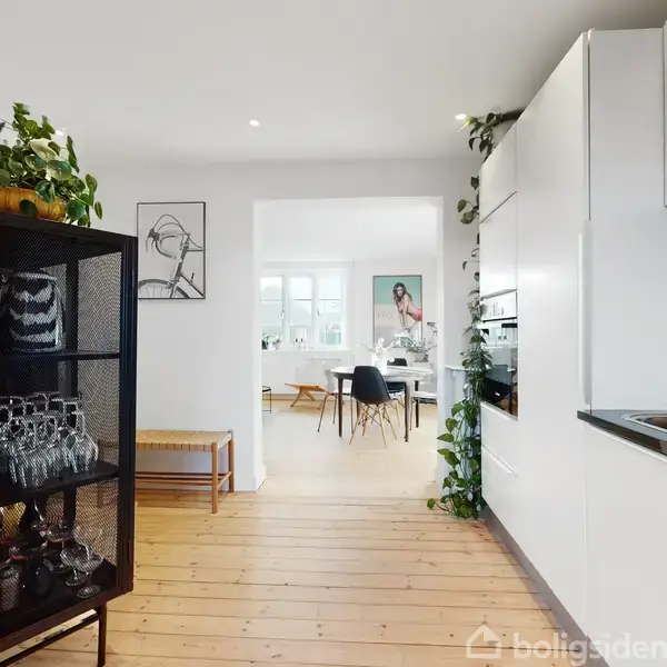 Hylde med glas, stående i et køkken med planter. I baggrunden ses en lys stue med billeder på væggen, trægulve og et bord med stole, der angiver en moderne indretning.