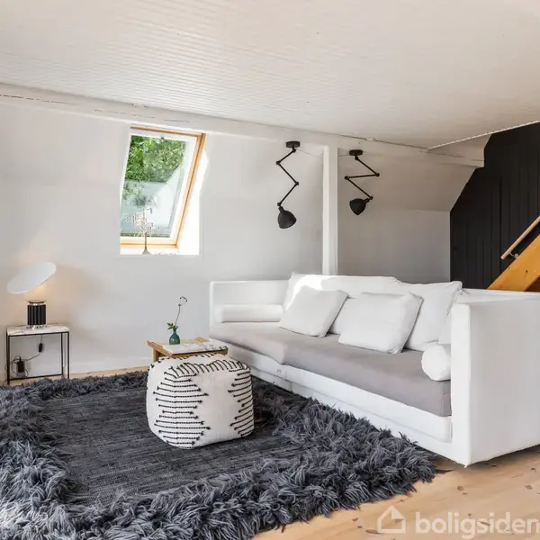 En hvid sofa står på et blødt, gråt tæppe. Et rundt, mønstret skammelbord er placeret foran sofaen. Trætrappen til højre fører til næste etage. En væg med et skråvindue lyser rummet op.