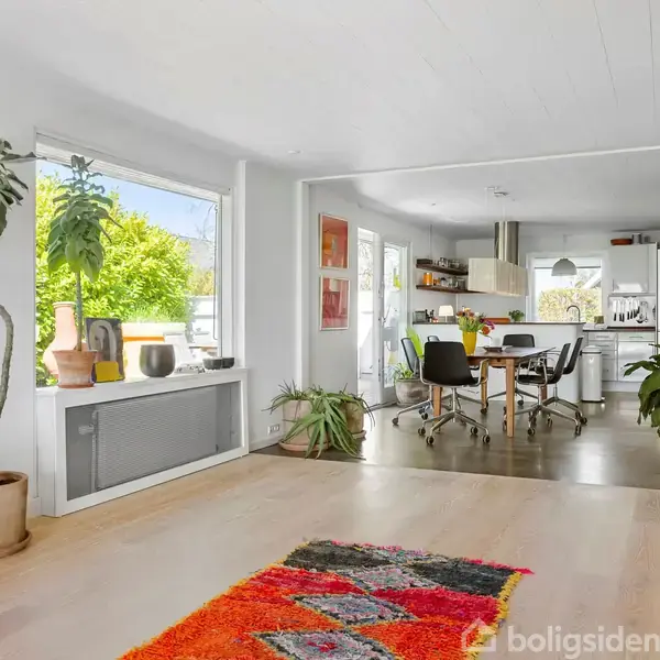 Et moderne hus med stor glasfacade står i en velplejet have. En terrasse med parasol rummer havemøbler, omgivet af farverige buske og træer. Blå himmel og solskin sætter scenen.