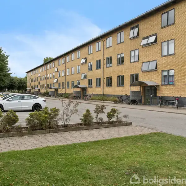 En gul murstensbygning med fire etager står langs en asfaltvej. En hvid bil er parkeret foran bygningen. Græsplæne med små buske omkranser en sti nær bygningen.