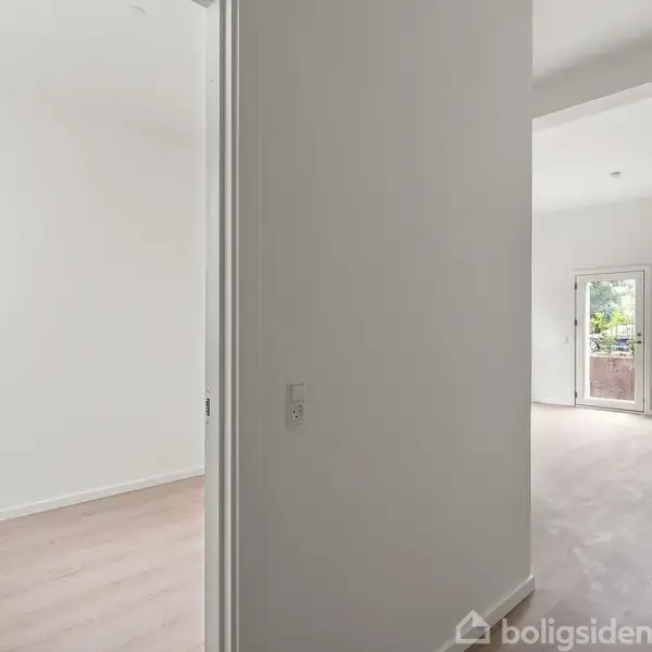 Døråbning fører til lyst rum med trægulv, hvidmalede vægge og dobbeltdøre, der åbner sig mod en balkon med grøn udsigt.