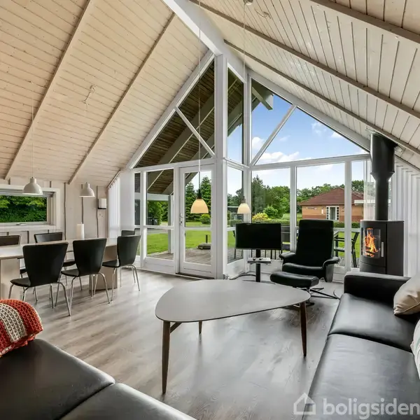 En stue med skrå loft, sorte lædersofaer, og et sofabord i træ. En brændeovn står nær vinduerne, der giver udsigt til en grøn have. Spisebord med stole er placeret ved vinduet.
