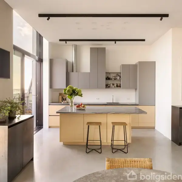 Køkkenø fungerer som samlingspunkt, flankeret af to taburetter. Moderne køkkendesign med gråt og lyst træ. Vaske og skabsplads i baggrunden. Lys strømmer ind fra store vinduer.