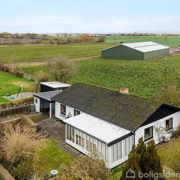 Et hvidt, énetages hus med sort tag ligger i en have med buske og træer. Ved siden af huset ses en fodboldbane. I baggrunden strækker marker sig mod horisonten.