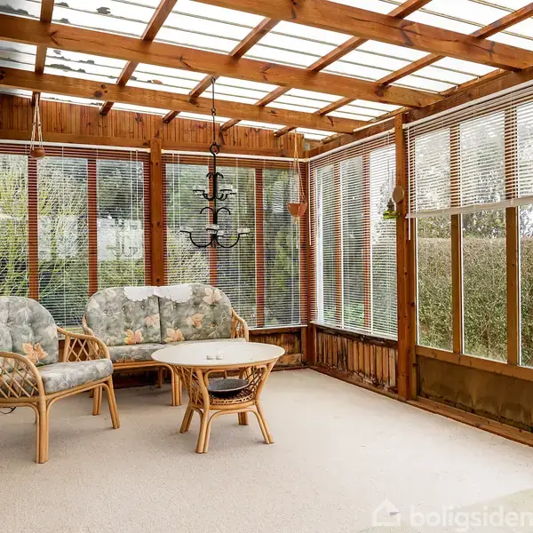 Fletmøbler, med blomstret betræk, står i en udestue. Et bord og stol er midtpunktet, omgivet af store vinduer og trærammer. En lysekrone hænger fra loftet.