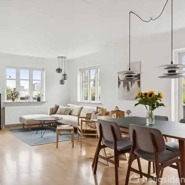 Stue med spisebord, der har solsikker i en vase, ved siden af en sofagruppe. Store vinduer giver naturligt lys. Lysekroner hænger fra loftet, skabe står langs væggen.