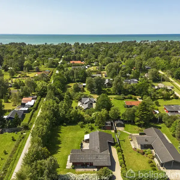 Luftfoto viser et kvarter med huse omgivet af grønne træer og græsplæner, der strækker sig mod horisonten, hvor havet kan ses.