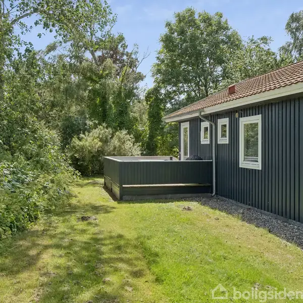 Et sort træhus med hvide vinduesrammer står stille ved en terrasse. Græsplæne og frodig skov omkranser huset, hvilket skaber en naturskøn og privat atmosfære.