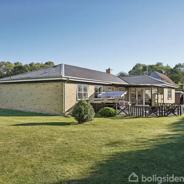 En gulstensvilla med sort tag er omgivet af en velholdt græsplæne. Der er en stor terrasse med havehegnet omkring. Huset ligger i et grønt, skovomkranset område.