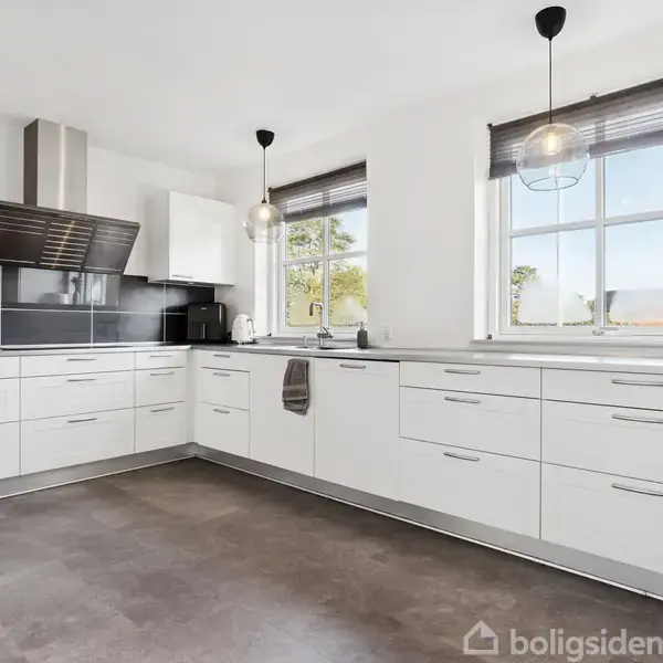 Lyst køkken med hvide skabe er indrettet i en U-form. Køkkenet har sort bordplade, hængende lamper, to vinduer med rullegardiner og mørkt gulv. Forskellige apparater og redskaber er synlige.