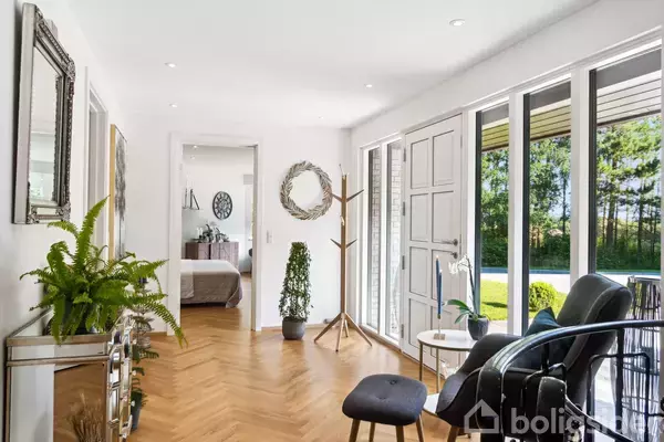 Entré med planter på et skrivebord mod venstre, åbne franske døre til en terrasse til højre. I baggrunden ses et soveværelse med seng.