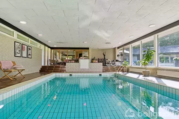 En indendørs swimmingpool står stille, omkredset af store vinduer, der lader naturligt lys strømme ind. Ved poolens ene side ses planter og møbler med hvide vægge og loft.