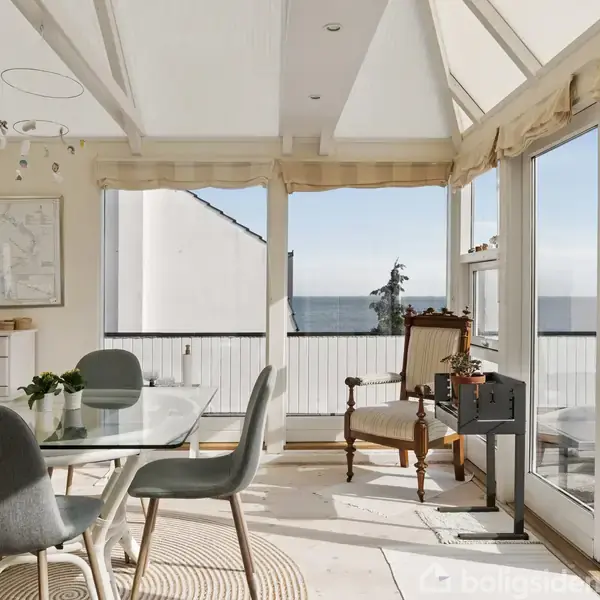 Et lyst rum med et glasbord omgivet af grå stole. Solen skinner ind fra store vinduer, der åbner ud mod en balkon med udsigt over havet.