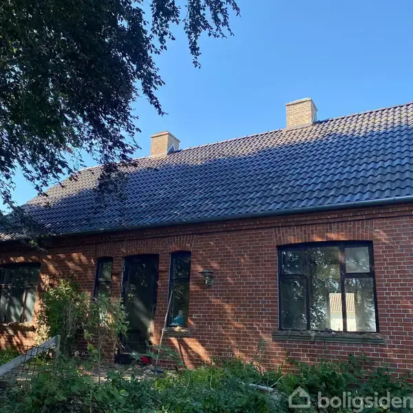Et rødt murstenshus med sort tegltag står omkranset af en have med træer og buske. Skyggen fra træerne falder på husets facade under en klar blå himmel.