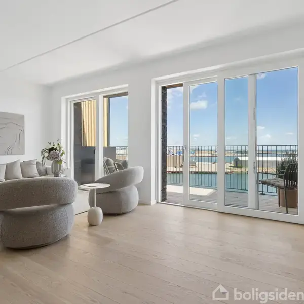 Lyst stueområde med moderne sofa og stole, vendt mod store vinduer, der åbner ud til en balkon med udsigt over vandet. Et kunstværk hænger på væggen. Lys trægulv.
