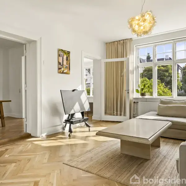Stue med stor beige sofa, sofabord og staffeli. Sollys strømmer ind fra vinduerne. I baggrunden ses en spisestue med trægulv og spisebord.