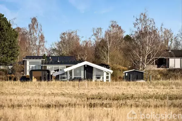 Et moderne, hvidt sommerhus står stille omgivet af høje træer og græs i en åben natur. Himlen er klar og blå, hvilket indikerer solskinsvejr.
