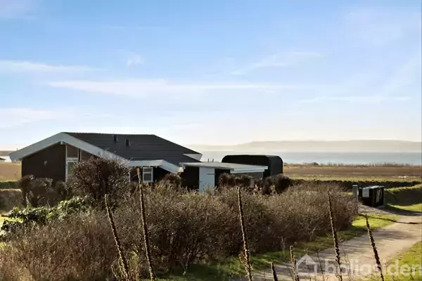 Et hus ligger stille omgivet af buske. Baggrunden viser en vidstrakt udsigt over en blå havhorisont og en klar himmel under sollys.