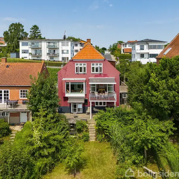 Rødt hus med hvidt tag står stille mellem to andre huse i et beboelsesområde, omgivet af grønne buske og træer, med en klar blå himmel i baggrunden.