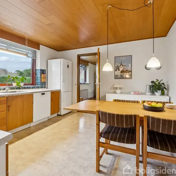 Et køkken med træloft har et spisebord med to stole, hængende lamper, og udsigt gennem et vindue. Der er køkkenskabe, en vask, en køleskab, og dekorationer på væggen.