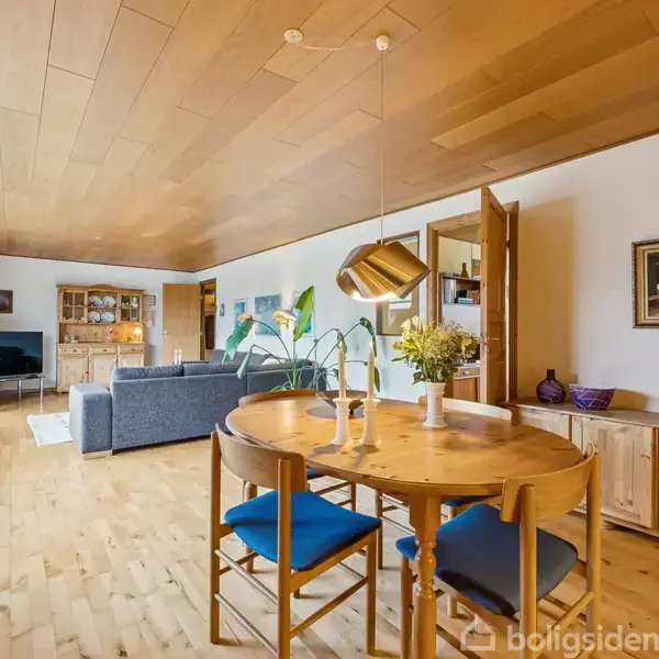 Et rundt spisebord med blå stole står i et lyst rum med trægulv og træloft. Baggrunden indeholder en sofa, store vinduer og diverse dekorative elementer.