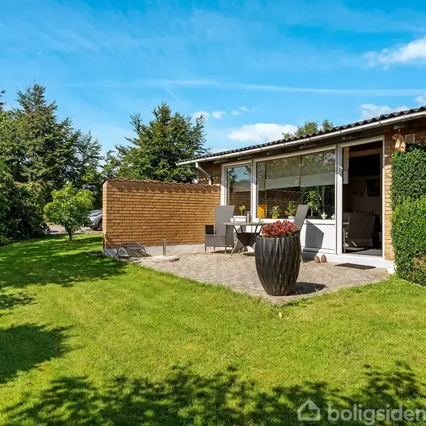 Et parcelhus med store vinduer åbner sig mod en terrasse, hvor havemøbler står. Græsplænen er omkranset af buske og træer, og himlen er klar og blå.
