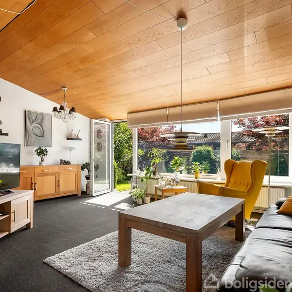 Stue med sort lædersofa, træbord, gul lænestol, fladskærms-tv og træloft. Åben dør viser frodig have omgivet af grønne planter. Møbler er moderne og lyset er indbydende.