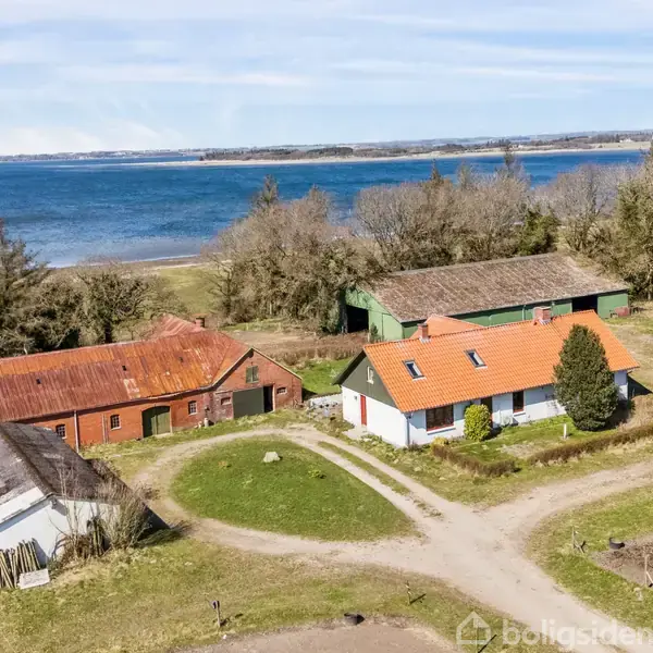 Et lille gårdkompleks med en hovedbygning og to udhuse ligger ved kysten. Hovedbygningen har et rødt tegltag. Baggrunden består af træer og havudsigt.