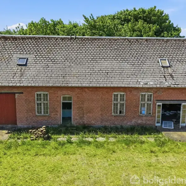 En rød murstenslade med skråtag og to loftvinduer står i en grønt landskab. Døre og vinduer er åbne. Baggrunden er træbevokset.
