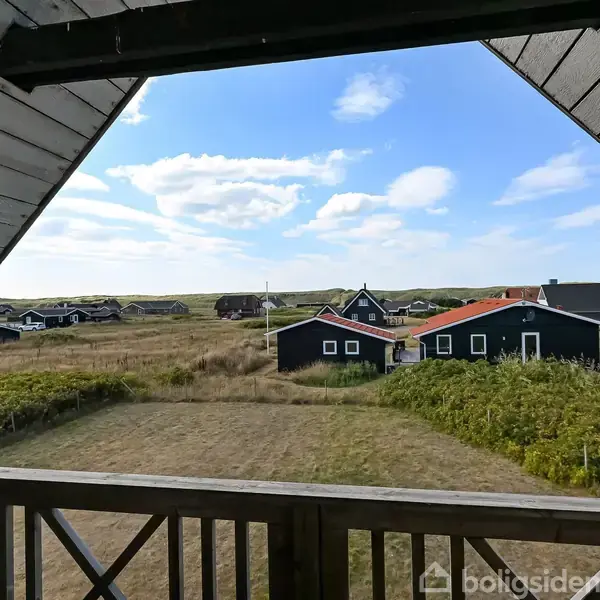 Balkon med udsigt over et åbent landskab med flere sorte, træhuse. Himmel med spredte skyer indrammer scenen. En dør står åben, og grønne buske gror omkring husene.