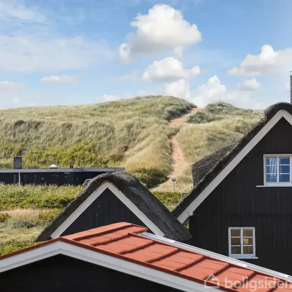 To sorte huse med stråtag står ved en sti, der fører op til en græsklædt klit under en blå himmel med hvide skyer.