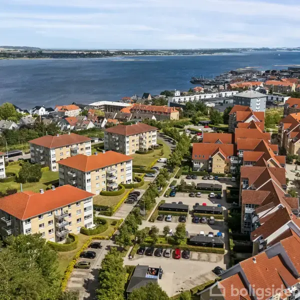 Flere bygninger med røde tage står spredt ud over et frodigt grønt boligområde ved en vandkyst. Vandet strækker sig mod horisonten, flankeret af byen i baggrunden.