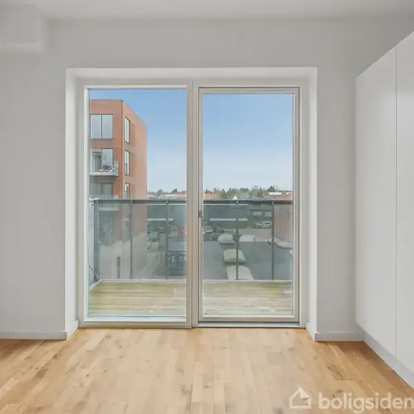 Glasdør åbner til en balkon med udsigt over en moderne bygning. Omgivet af et lyst rum med trægulv og hvide vægge.