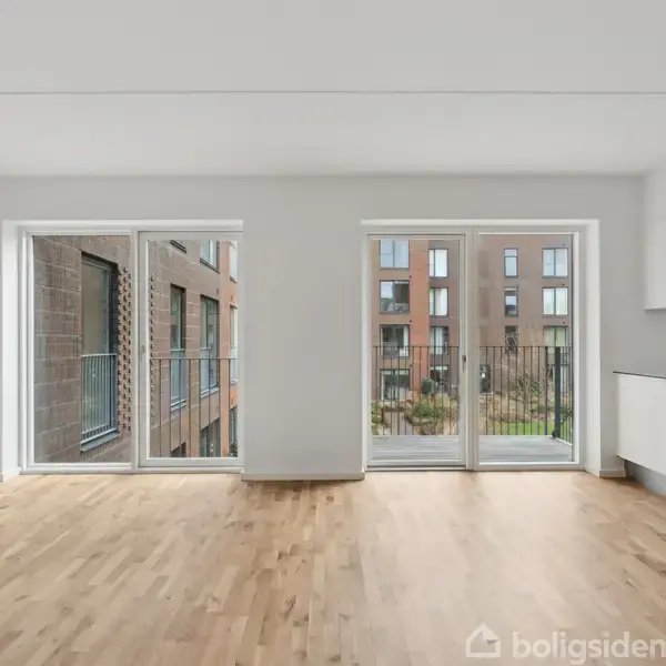 Et lyst, tomt rum med trægulv, store glasdøre åbner til en balkon med udsigt til en moderne bygning. Til højre er et hvidt, indbygget køkken med ovn og skabe.