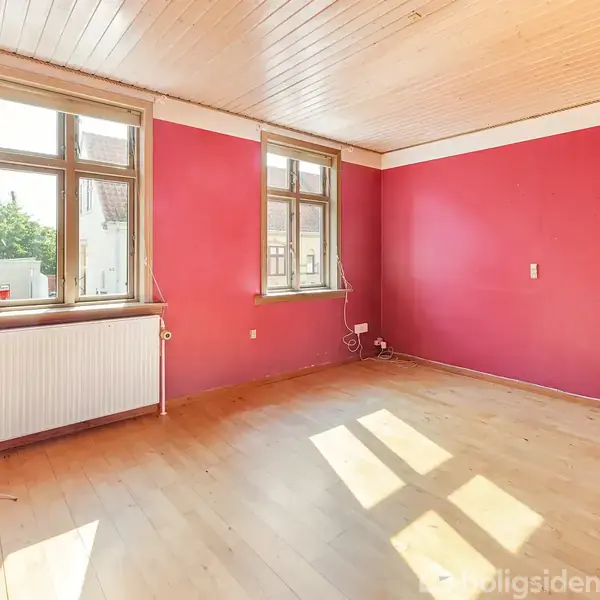 Rummeligt værelse med røde vægge og lyst trægulv. To store vinduer lader sollys strømme ind. Radiator under vinduerne, ingen møbler. Hvidt loft med træpaneler giver et åbent indtryk.