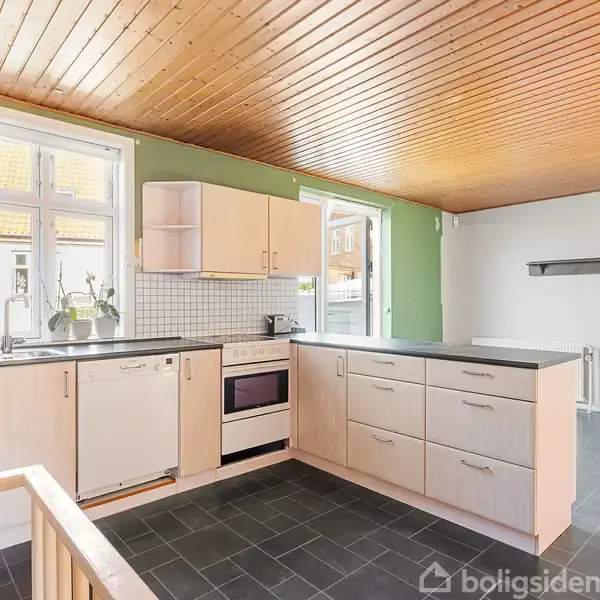 Et køkken med træloft og hvide skabe står tomt. Der er en ovn, en vask, og et vindue, der lukker dagslys ind. Boligen er lys med grønne vægge.