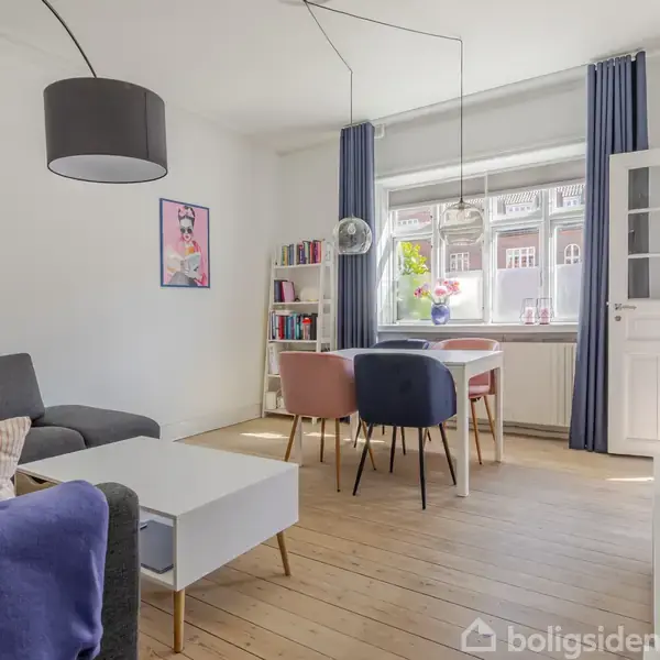 Stue med en grå sofa, sofabord og et spisebord med farverige stole. Store vinduer med blå gardiner lader lys ind. En bogreol og vægkunst pryder rummet.