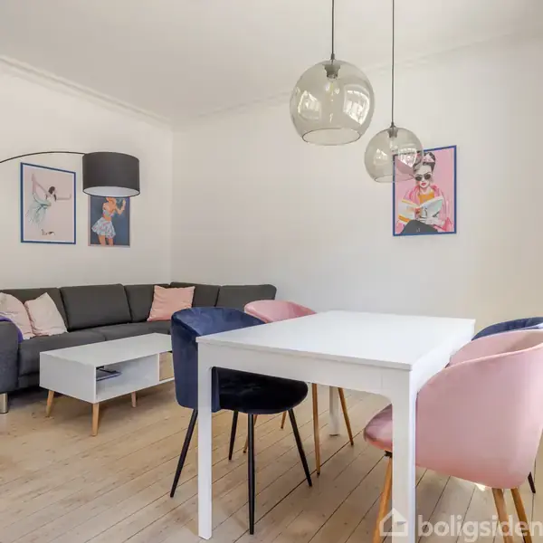 Gråt hjørnesofa står i en lys stue, dekoreret med farverige puder og to kunstplakater. Tre hængelamper hænger over en hvid spisebord med forskellige farvede stole. Indretning er moderne.