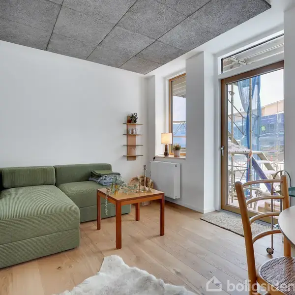En grøn sofa står i en lys stue med et sofabord foran. Der er en udgang til en balkon gennem en glasdør, og rummet har trægulv og et lille rundt bord.