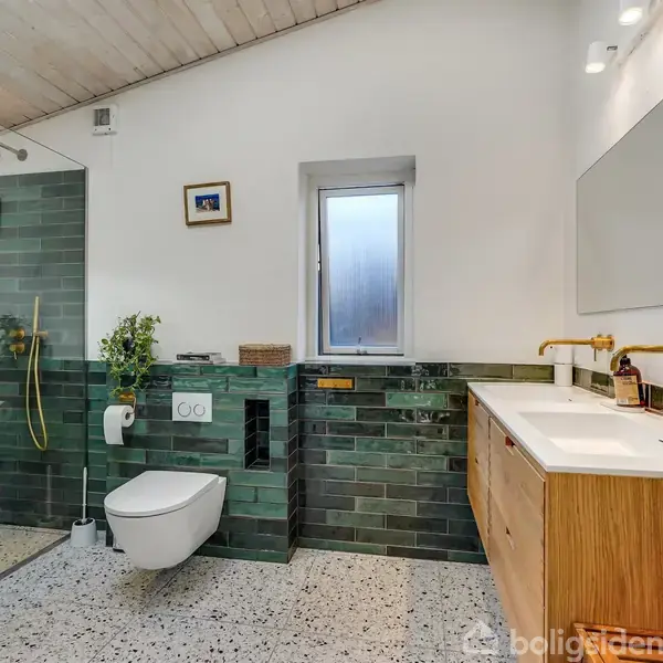 Badeværelse med grønne fliser og glasvæg omkranser bruseområdet. Til venstre hænger toilet. Til højre står en håndvask med træskab under et stort spejl. Et lille vindue centralt.