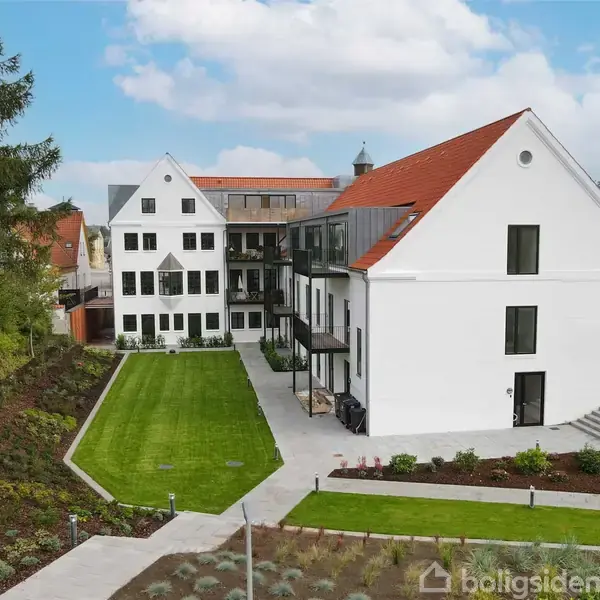 En større hvid bygning med rødt tag står stille i en velplejet have med græsplæne og stier. Omkringliggende træer og en blå himmel skaber en fredelig atmosfære.