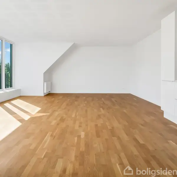 Et lyst tomt rum med trægulv; store vinduer lader sollys ind. Ved siden af, er der en hvid væg med indbyggede skabe. Udsigt over grønne træer udenfor.