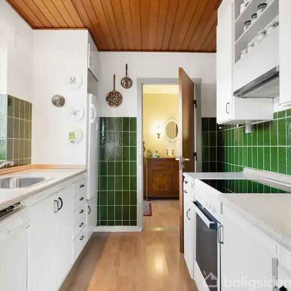 Et lyst køkken med hvide skabe og grønne fliser. Vasken og opvaskemaskinen står ved vinduet. I baggrunden ses en åben dør til et andet rum med et spejl og et skab.