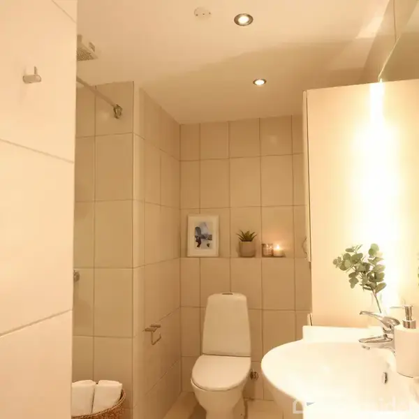 Toilet står stille i et lyst, flisebelagt badeværelse med brusekabine. På væggen hænger et billede, og ved spejlet står planter og stearinlys, der tilføjer en hyggelig atmosfære.