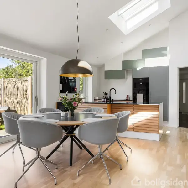 Rundt spisebord med fire grå stole står under en stor lampe i et lyst køkken-alrum. Store vinduer åbner ud til terrassen med grøn udsigt. Skylight ses i loftet.