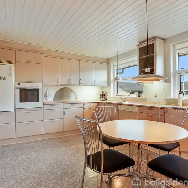 Et køkken med lyse træskabe langs væggen. Et rundt spisebord med fire stole står centralt. To store vinduer giver udsigt til et boligkvarter.